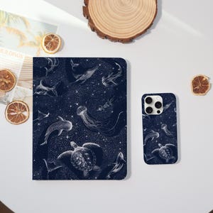 Op de afbeelding: Een donkerblauwe notitieboek en telefoonhoesje met een witte illustratie van verschillende zeedieren, waaronder walvissen, haaien en kwallen, tegen een sterrenachtergrond. Het notitieboek en de telefoonhoesje liggen op een wit oppervlak.