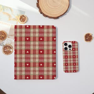 May include: A red and beige plaid pattern notebook and phone case with white heart accents. The notebook is rectangular, and the phone case has a camera cutout. The items are displayed on a white surface with dried orange slices and a wooden coaster.