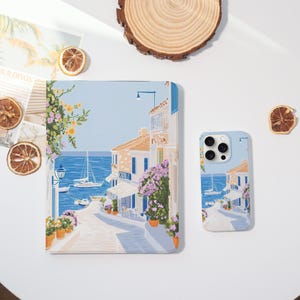 Può includere: Una custodia per telefono e una custodia per tablet abbinate con un design di città costiera. Le custodie raffigurano una scena di strada con edifici, barche e fiori, su uno sfondo azzurro. Le custodie sono illustrate.