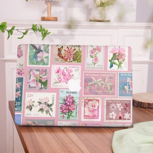 May include: A pink laptop case adorned with a collage of vintage postage stamps featuring floral designs and botanical illustrations. The stamps showcase various countries and denominations, creating a unique and artistic aesthetic. The case has a smooth finish.