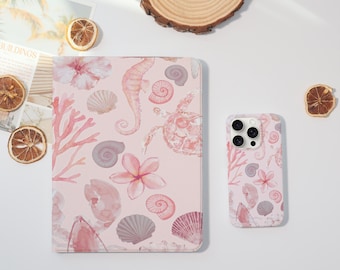 Può includere: Una custodia rosa per tablet e telefono con un design ad acquerello con cavallucci marini, conchiglie, coralli e fiori. Le custodie sono su una superficie bianca con fette d'arancia essiccate e un sottobicchiere di legno.