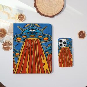 Peut inclure: Un design pop art bleu et orange représentant un OVNI avec un œil sur le dessus, émettant un flux orange et jaune. Le design est sur une coque de tablette et une coque de téléphone.