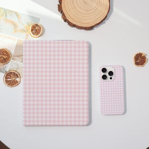 Peut inclure: Une housse de tablette et une coque de téléphone à motif vichy rose et blanc. Les étuis sont sur une surface blanche avec des tranches d'orange séchées et un dessous de verre en bois. L'étui de la tablette est rectangulaire.