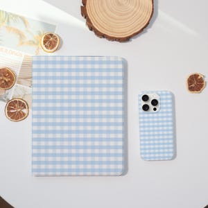 May include: A light blue and white gingham pattern notebook and phone case. The notebook is closed and rectangular. The phone case has a camera and flash. The items are on a white surface.