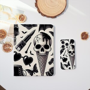 Op de afbeelding: Een crèmekleurige tablethoes en telefoonhoesje met een zwart-wit schedel-ijshoorntjesontwerp. Het ontwerp bevat vleermuizen, lippenstift, harten en rozen. De hoesjes liggen op een wit oppervlak.