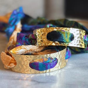 May include: Two gold-colored, hammered metal bracelets with oval cutouts, each holding a colorful fabric. The fabric is a mix of blue, purple, and yellow. The bracelets are stacked on top of each other.