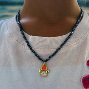 May include: A beaded necklace with a gold-colored paw print pendant featuring a red heart. The pendant is encrusted with small, clear stones. The necklace is worn over a white and pink striped t-shirt with a red embroidered design.