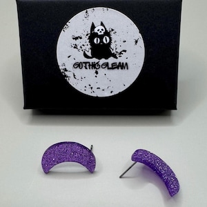 May include: A pair of purple crescent moon shaped earrings with glitter. The earrings are packaged in a black box with a white sticker that says "Gothic Gleam" and has a black cat with a skull on its head.
