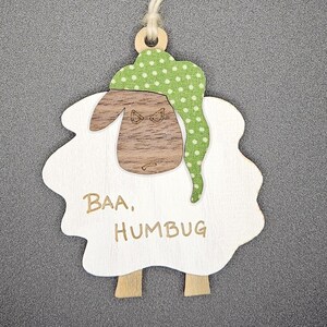 May include: A wooden sheep ornament with a white body, brown face, and green polka-dot hat. The text "Baa, Humbug" is printed on the sheep. A natural twine string is attached for hanging. The ornament is designed for the holiday season.