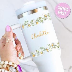 May include: White travel mug with a handle, featuring a daisy floral design and the name "Charlotte" in gold lettering. The mug has a white lid and a silver band near the top. A pink sticker with "SHIPS FAST" is visible.