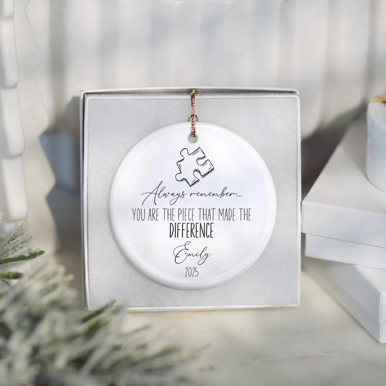May include: A white ceramic ornament featuring a puzzle piece design and the inscription "Always remember... You are the piece that made the difference Emily 2025." The ornament is presented in a white gift box.
