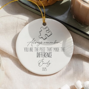 May include: Round white ceramic ornament with a gold hanging string. It has a puzzle piece design and the inscription: "Always remember... YOU ARE THE PIECE THAT MADE THE DIFFERENCE Emily 2025."