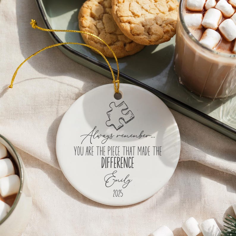 May include: A round white ceramic Christmas ornament with a gold hanging string. The ornament has a puzzle piece design and the text: "Always remember... YOU ARE THE PIECE THAT MADE THE DIFFERENCE Emily 2025."