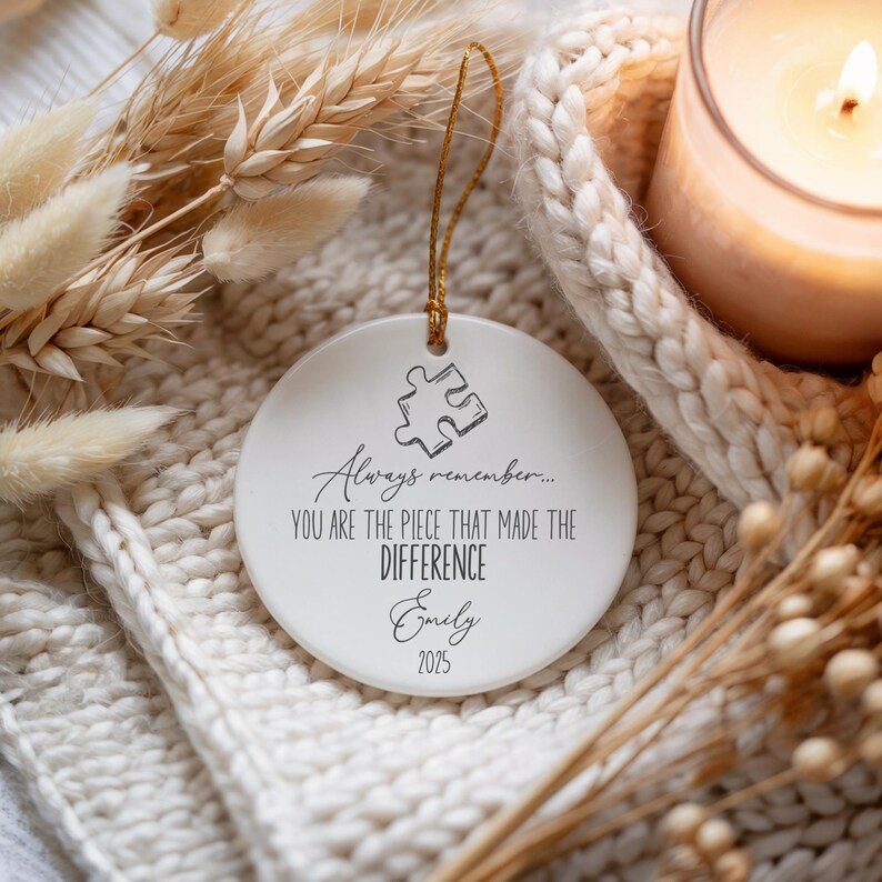 May include: A white ceramic disc ornament with a gold hanging string. It displays a puzzle piece graphic and the text "Always remember... YOU ARE THE PIECE THAT MADE THE DIFFERENCE Emily 2025". The ornament is set amongst dried flowers and a lit candle.