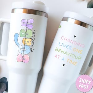 May include: Two white insulated tumblers with handles. One features the name "Amelia" and the text "BOARD CERTIFIED ASSISTANT BEHAVIOUR ANALYST" in pastel colors. The other tumbler has the text "CHANGING LIVES ONE BEHAVIOUR AT A TIME" in pastel colors.