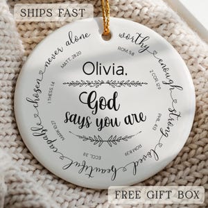 May include: A white ceramic ornament with a gold string for hanging. The ornament features the name "Olivia" and the phrase "God says you are" in black script. Surrounding the text are words like "chosen" and "beautiful".
