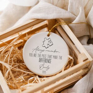 May include: A white ceramic ornament with a puzzle piece design and the text "Always remember... YOU ARE THE PIECE THAT MADE THE DIFFERENCE Emily 2025". The ornament is in a wooden crate with straw.