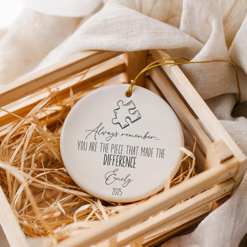 May include: A white ceramic ornament with a puzzle piece design and the text "Always remember... YOU ARE THE PIECE THAT MADE THE DIFFERENCE Emily 2025". The ornament is in a wooden crate with straw.
