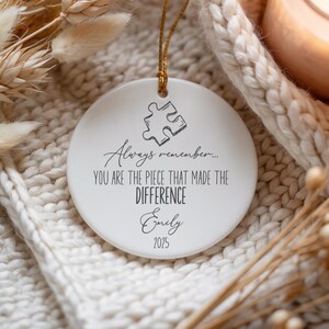May include: A white ceramic ornament with a gold string. The ornament has a puzzle piece graphic and the text "Always remember... YOU ARE THE PIECE THAT MADE THE DIFFERENCE Emily 2025". It is displayed on a white knitted surface.