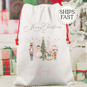 May include: White Christmas gift bag with red drawstring closure. The bag features a festive illustration of a Christmas tree, nutcrackers, and a ballerina. The words "Merry Christmas to" and a name are printed on the bag.