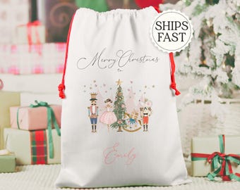May include: White Christmas gift bag with red drawstring closure. The bag features a festive illustration of a Christmas tree, nutcrackers, and a ballerina. The words "Merry Christmas to" and a name are printed on the bag.