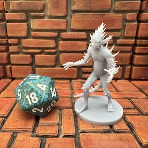 May include: A gray, spiky fantasy figure stands on a circular base next to a large, blue-green twenty-sided die. The figure has a humanoid form with sharp protrusions. The die is numbered with white numerals.