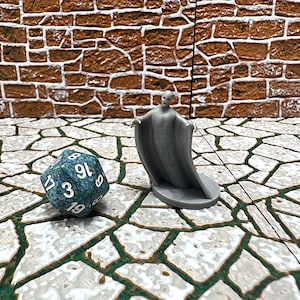 May include: A gray miniature ghost figure with outstretched arms stands next to a blue and green 20-sided die. The die has white numbers. The background features a brick wall and a stone-like surface.