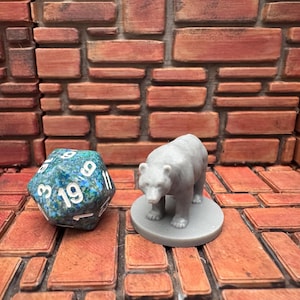 May include: A gray bear figurine on a gray base stands next to a large, multi-sided die with white numbers on a teal and blue surface. The background is a brick-like pattern in shades of brown and orange.