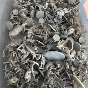 Puede incluir: Una gran colección de figuras en miniatura de plástico gris, algunas con armas, en un contenedor de plástico gris. Las figuras están sobre bases redondas.