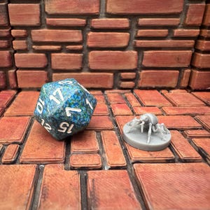 Pode incluir: Um dado de vinte lados salpicado de azul e verde com números brancos, ao lado de uma pequena figura de aranha cinzenta. Os itens estão em uma superfície semelhante a tijolos com um fundo de parede de tijolos. O dado é usado para jogos de tabuleiro.