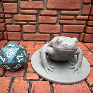 May include: A gray, three-dimensional frog figurine on a round base, next to a blue and green polyhedral die. The die has white numbers and is a 20-sided shape. The background is a brick wall.