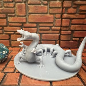 May include: A light grey, 3D-printed monster figurine with an open mouth and multiple legs, posed on a circular base. A multi-sided die with blue and green hues is next to the figurine. The background is a brick wall.
