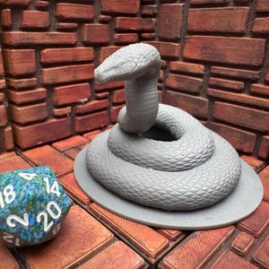 Op de afbeelding: Een grijze, opgerolde slangenfiguur met opgetild hoofd, op een cirkelvormige basis. Een veelzijdige dobbelsteen met blauwe en groene patronen en witte cijfers staat in de buurt. De achtergrond heeft een baksteenachtig patroon.