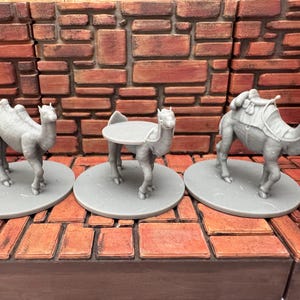 May include: Three gray camel figurines on round bases. One camel stands alone, another has a tabletop, and the third has a saddle and pack. The figurines are set against a brick wall backdrop.