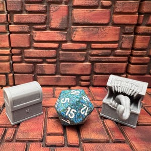 May include: Three gray miniature treasure chests and a blue and green twenty-sided die with white numbers are displayed on a brick-like surface. One chest is closed, another open with a tongue, and the die shows the number 15.