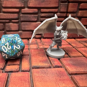 May include: A blue and green twenty-sided die with white numbers sits next to a gray gargoyle figurine holding a skull. The gargoyle has large wings and is on a round base. The background is a brick-like surface.