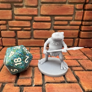 May include: A gray frog-like miniature figure holding a spear, standing on a circular base. A multi-sided die with blue and green patterns and white numbers is next to the figure. The background is a brick wall.