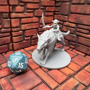 May include: A gray miniature figure of a goblin riding a wolf-like creature, holding a sword. A blue and green 20-sided die is next to the figures. The figures are on a gray base, set against a brick-like background.