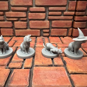 Pode incluir: Quatro estatuetas de gato cinzentas sobre bases redondas. Um gato usa um cachecol, outro uma venda, um terceiro está em pose de perseguição e o último usa um chapéu de bruxa. As estatuetas estão sobre uma superfície semelhante a tijolos.
