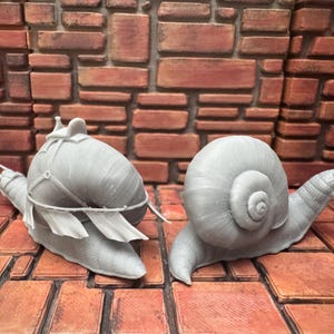 May include: Two gray snail figurines. One snail has a saddle and decorative elements, while the other is a simple snail. The snails are on a brick-patterned surface with a brick wall background.