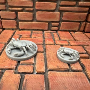 May include: Two gray crab-shaped miniatures on a brick-patterned surface. One is larger, the other smaller, both with detailed crab features. The background is a brick wall. These miniatures are likely for tabletop gaming or crafting.