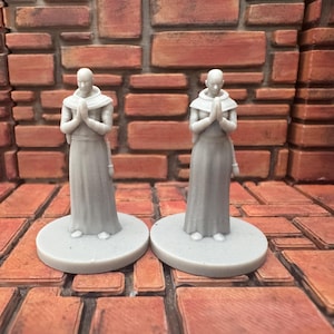 May include: Two gray figurines of figures in robes with hands clasped in prayer. The figures stand on round bases. The background is a brick wall and brick floor. The figures are approximately 4 inches tall.