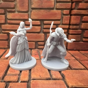 May include: Two gray miniature fantasy figures. One figure is a woman in a long dress, holding a book and raising her hand. The other figure is a man with a staff and cape, reaching out. Both figures are on round bases.