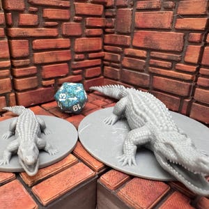 May include: Two gray alligator figurines, one large and one small, rest on gray circular platforms. A multi-sided blue and green die is between them. The background is a brick wall.