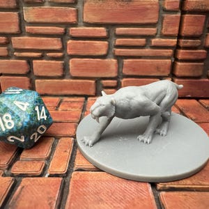 May include: A grey, 3D-printed sabre-toothed tiger figurine on a grey circular base, in a predatory pose. A multi-sided blue and green die with white numbers is to the left. The background is a brick wall.