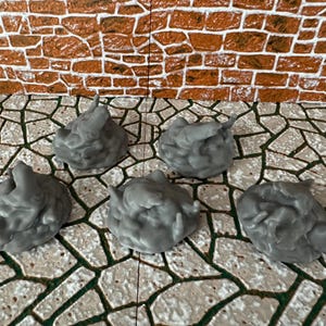 Può includere: Cinque miniature grigie a forma di nuvola con superfici strutturate. Questi piccoli modelli dettagliati sono probabilmente per giochi da tavolo o scenografie di modelli. Lo sfondo presenta un muro di mattoni e una superficie simile alla pietra.