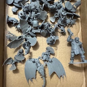 May include: A collection of unpainted grey plastic miniature figures, including various fantasy creatures, winged beings, and a large humanoid figure holding a club, all scattered together in a cardboard container.