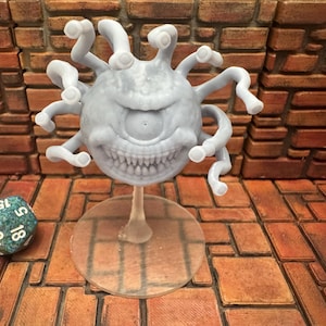 May include: A gray, 3D-printed beholder miniature with multiple eye stalks and a large central eye. The creature has a toothy grin and is mounted on a clear stand. A multi-sided die rests nearby, suggesting a tabletop game theme.