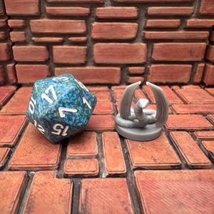May include: A blue and green speckled D20 die with white numbers next to a gray miniature serpent with wings. The objects are on a brick-patterned surface with a brick wall background. The items are for tabletop gaming.