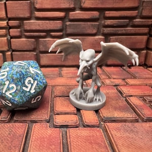 May include: A gray gargoyle figurine with outstretched wings stands next to a large, multi-sided die. The die is blue and green with white numbers. The background is a brick-like surface.